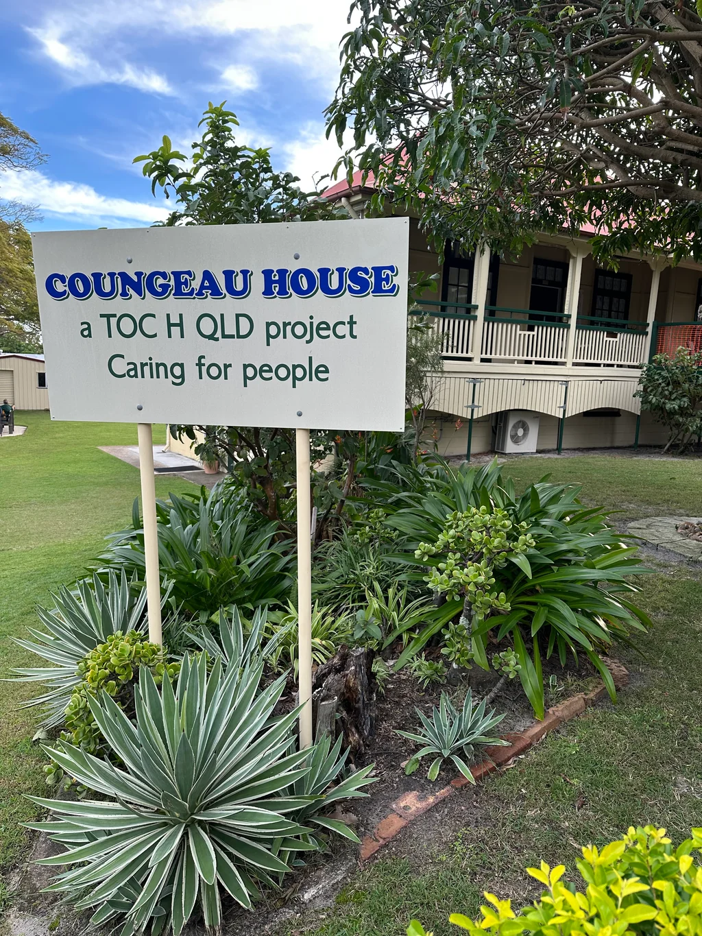 The Coungeau House sign reading 'a Toc H QLD project — Caring for people', with the heritage building in the background framed by tropical garden