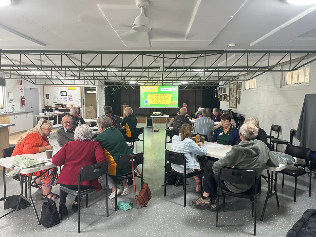 Members seated at tables across the hall during the Toc H trivia round, with a question on the projector screen