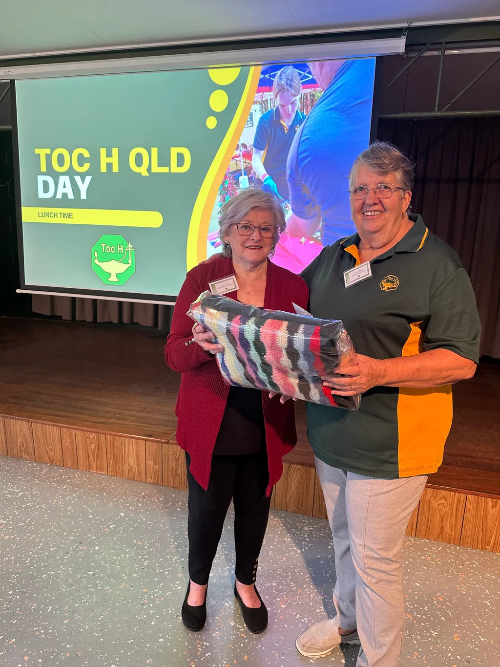 A handmade donation being presented to Bribie Branch in memory of a long-standing member, in front of the Toc H QLD Day screen